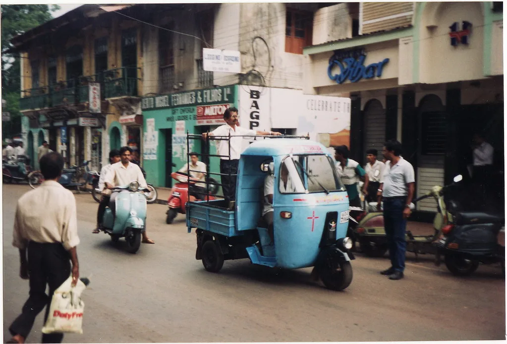 A driver at the wheel of a light-blue three-wheeled mini truck on a busy street in Goa