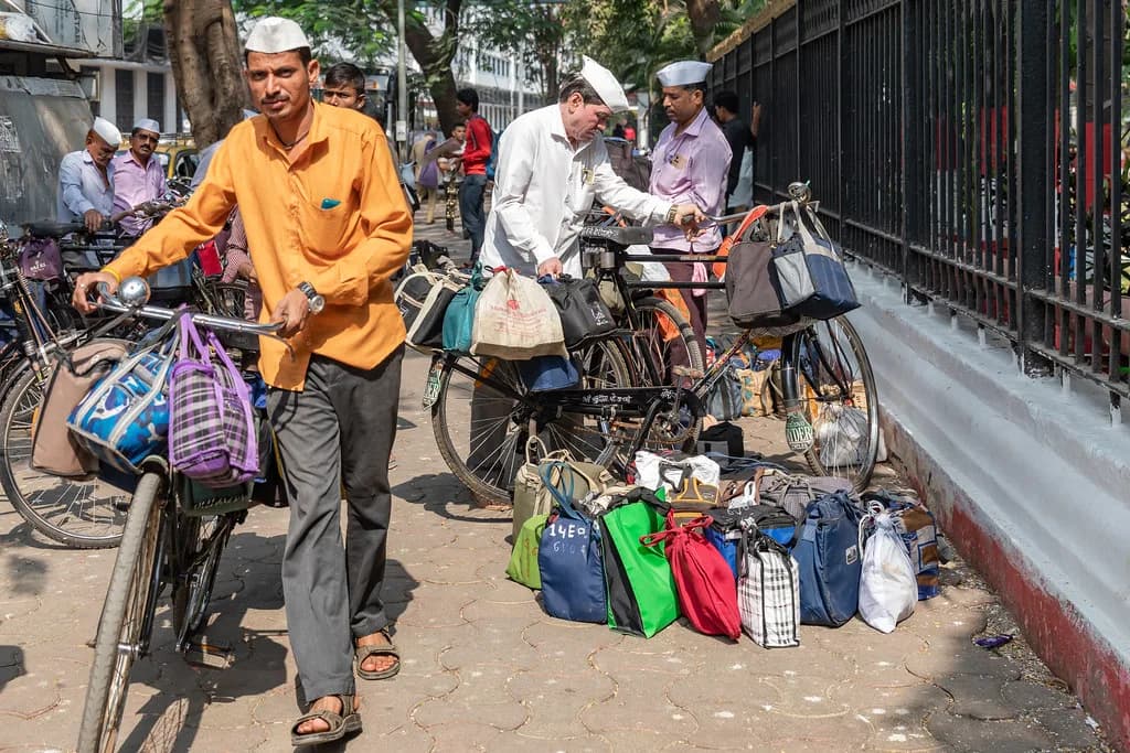 A dabbawala in a white cap wheeling a bicycle hung with tiffin bags past a row of loaded bicycles in Mumbai