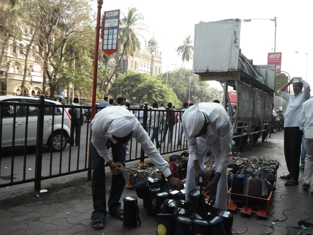 Dabbawalas bending over rows of tiffin bags on a Mumbai pavement, sorting them by destination
