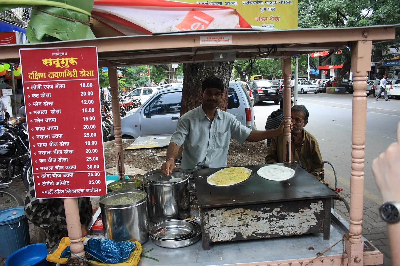 A cook making dosas on a griddle at a roadside stand, with a menu board beside him