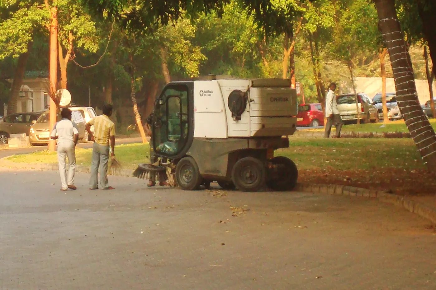 Two workers beside a compact sweeping machine on a tree-lined estate road
