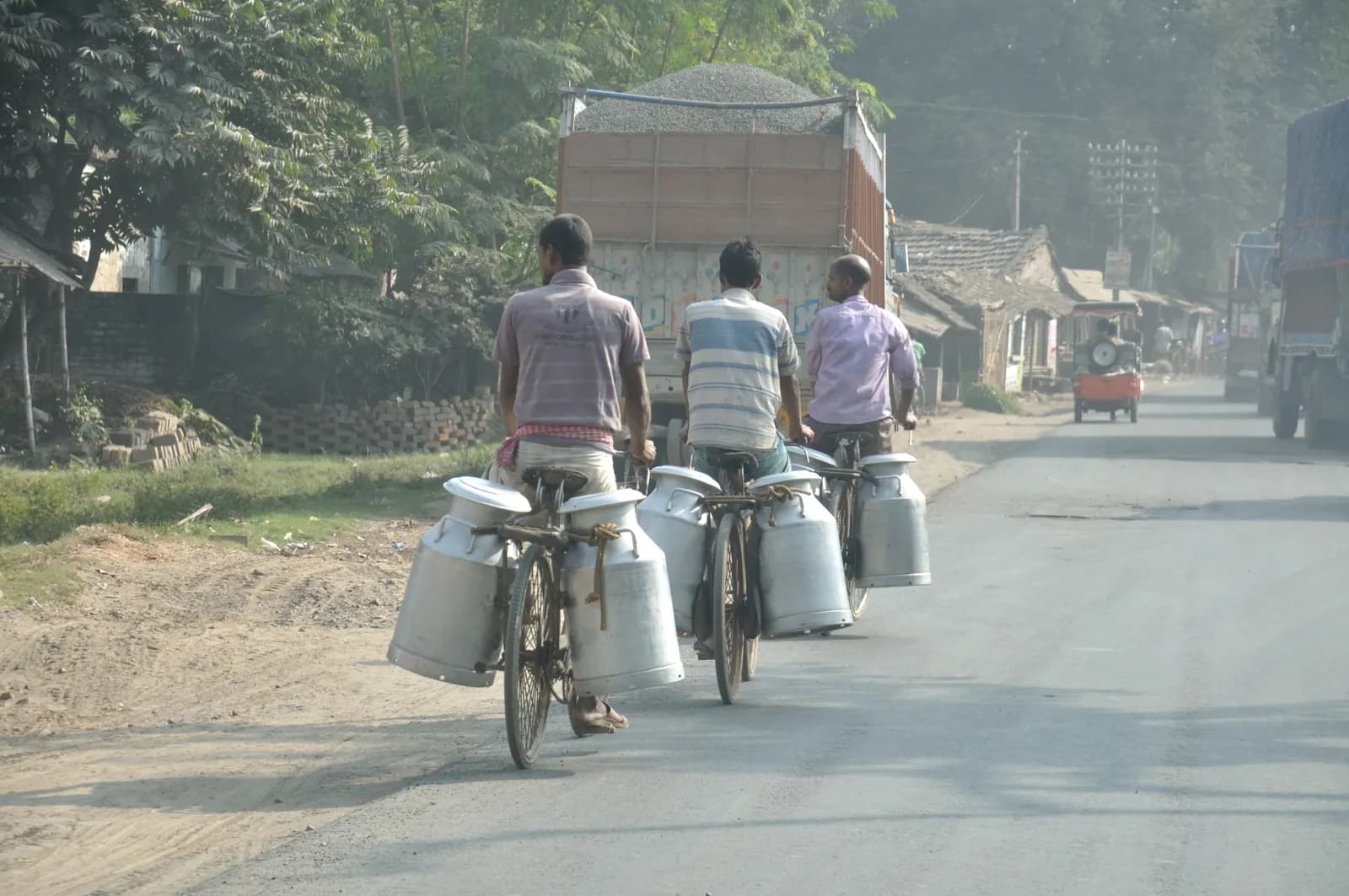 Three milkmen riding bicycles hung with large steel milk cans along a highway in West Bengal