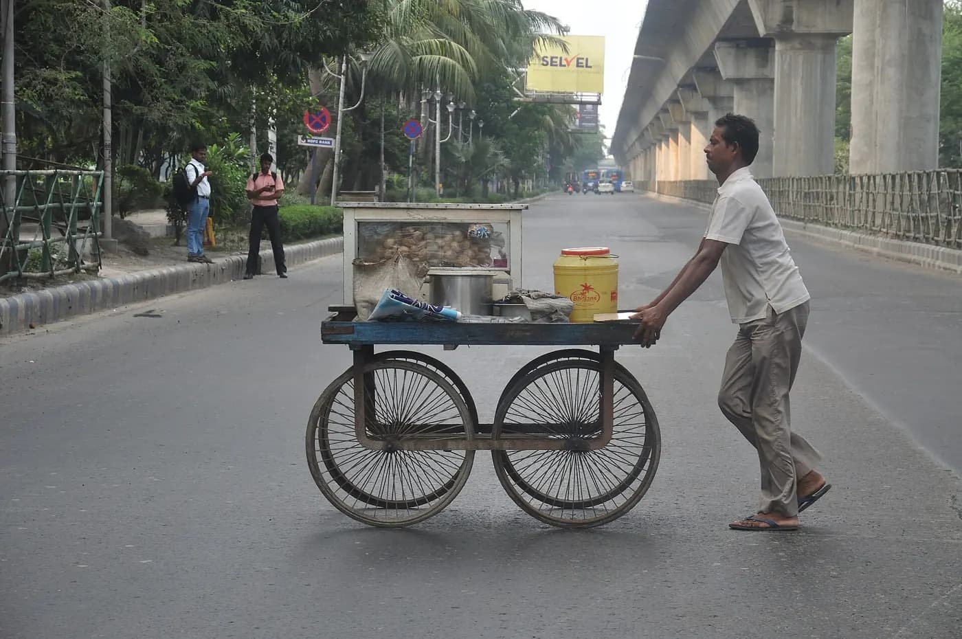 A pani-puri vendor pushing a loaded hand cart along a wide road