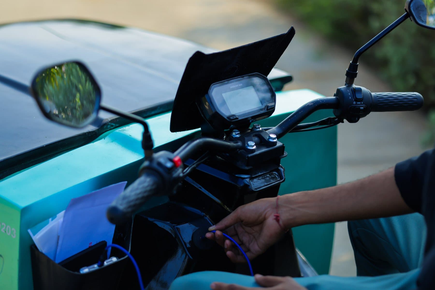 Close-up of a CROC EV's handlebar, mirrors and display, with a rider's hand connecting a small cable beside the teal cargo box.