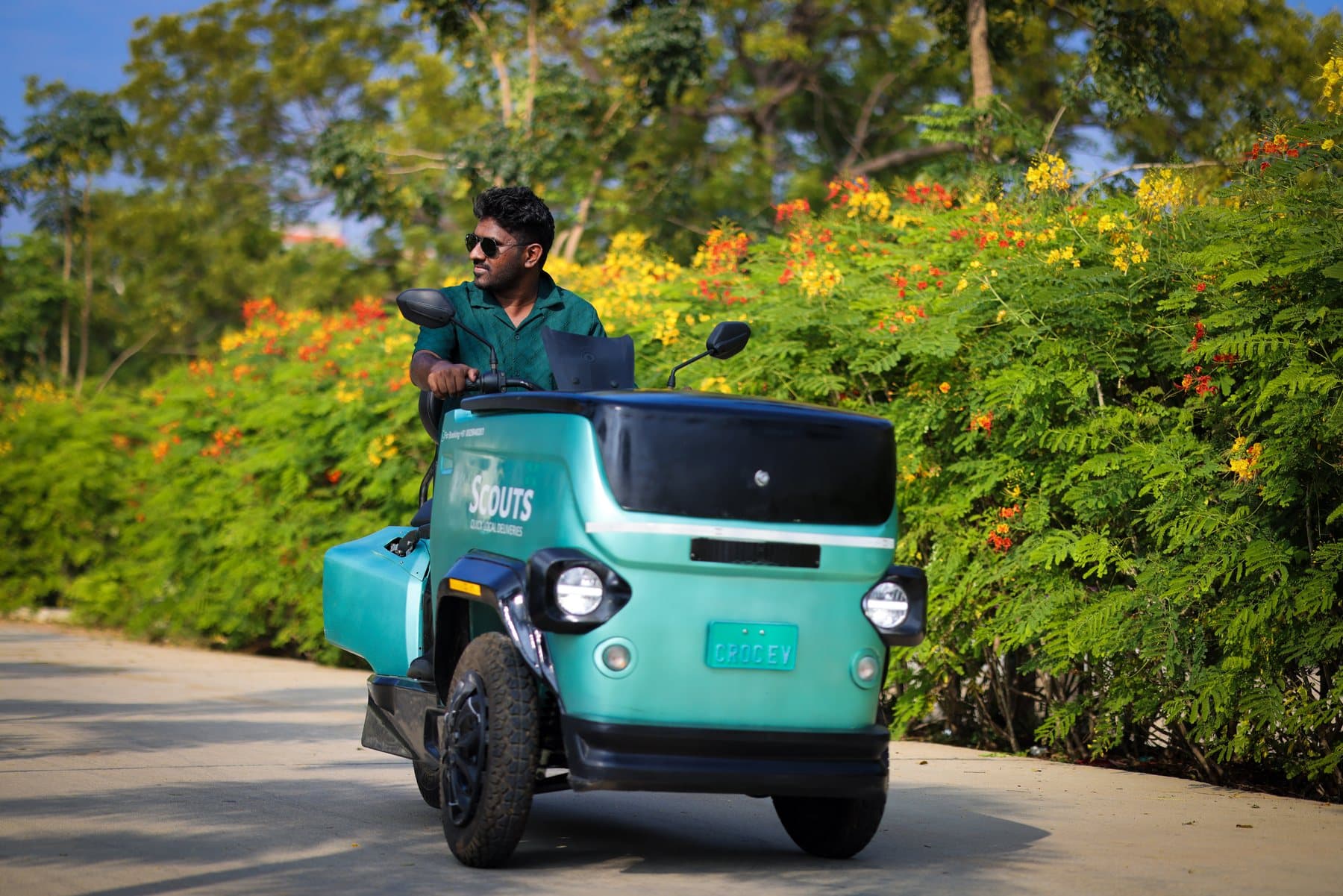 A rider steers a teal CROC EV along a tree-lined road, with its two front wheels and closed cargo box facing the camera.